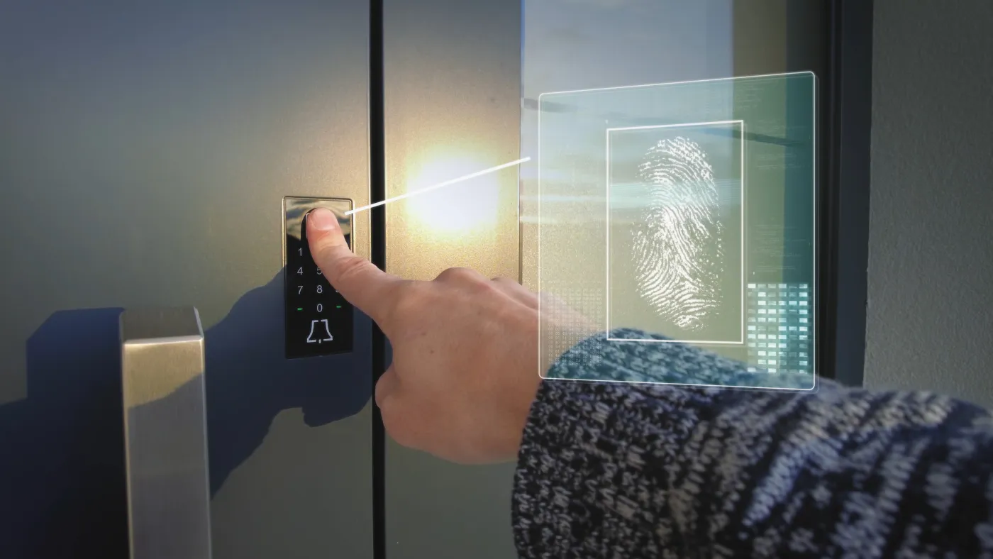 A finger on a backlit door keypad with a biometric fingerprint-scan overlay in warm evening light