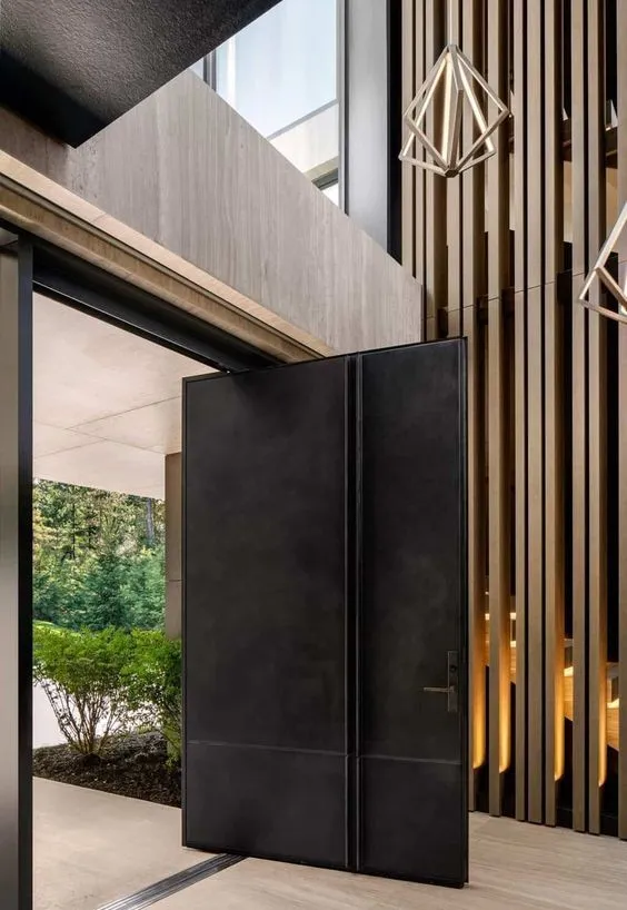 Black patinated-metal pivot door seen from a double-height atrium with a timber-slat screen and brass pendants