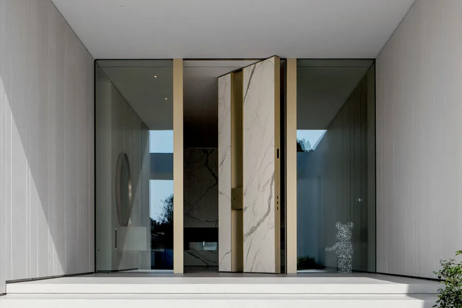 White marble-effect pivot door with brass vertical frame accents in a glass-walled minimalist portico