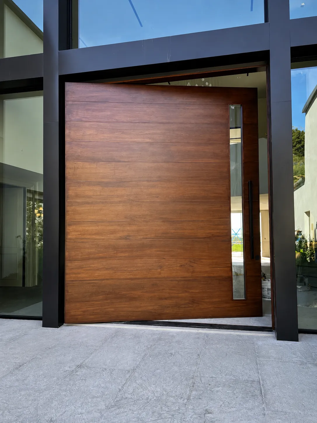 Oversized walnut aluminium pivot front door beside a full-height glass wall in daylight